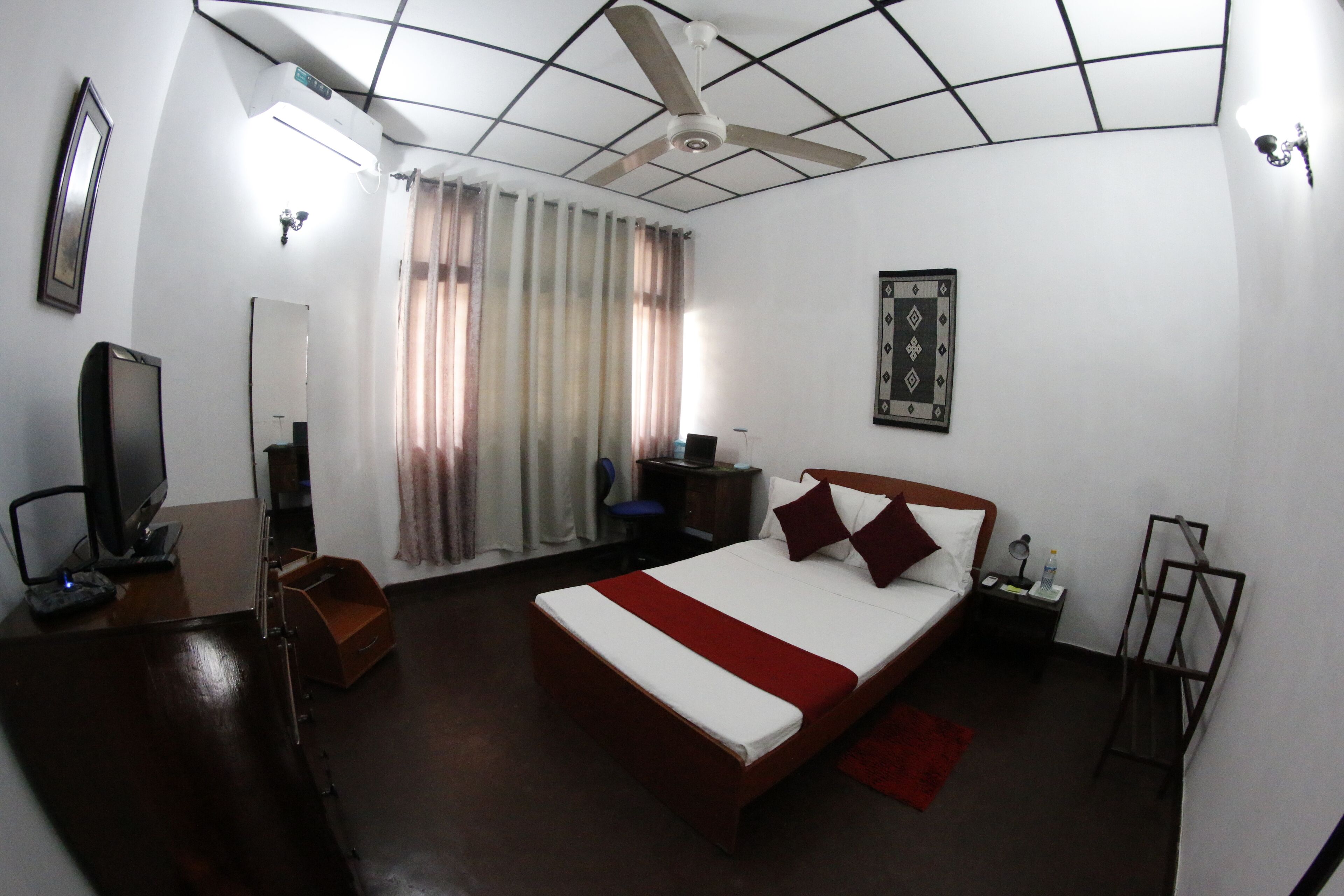 Habitación de hotel con cama, escritorio con computadora, televisor, ventilador de techo y ventana con cortinas.