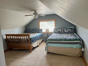 A bedroom with a sloped ceiling, a ceiling fan, a window with blinds, two beds with patterned bedding, a wooden crib, and a bedside table with a lamp.