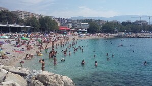 Ein überfüllter Strand mit badenden und sonnenden Menschen, felsiger Küstenlinie und Gebäuden im Hintergrund.