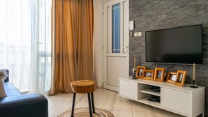 Ein Wohnzimmer mit einem großen Flachbildfernseher an einer Steinwand, einer weißen Unterhaltungsanlage mit Bilderrahmen, einem runden Holzschemel und einem großen Fenster mit Vorhängen.