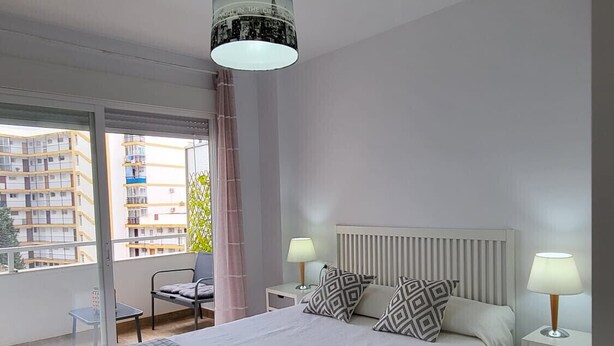 A modern bedroom with a large bed, two bedside tables with lamps, a view of buildings through a sliding door, and a hanging light fixture. - TERRASOL ESTRELLA 6 (Torre del Mar)