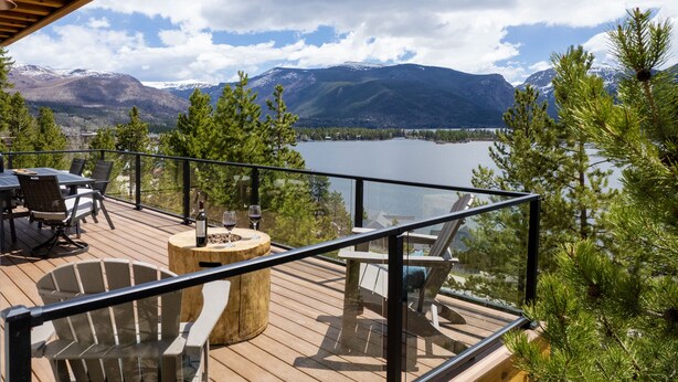 A deck with outdoor furniture, a round table with a bottle and glass, and a view of a lake and mountains. - Stunning Lake Views near RMNP with Hot Tub (Grand Lake)