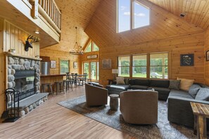 A spacious lodge interior with a stone fireplace, wooden ceiling, and large windows offering views of the outdoors.