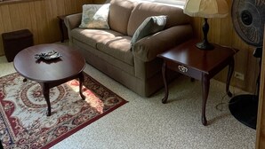 A cozy living room with a brown sofa, a small table, a lamp, and a fan.