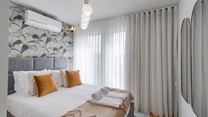 A hotel room with a bed, a large window with curtains, an air conditioning unit, and a wall with a floral pattern.