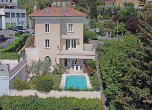A two-story building with a swimming pool, surrounded by greenery and adjacent to a road. - Villa San Carlo - Gardagate (Roè)