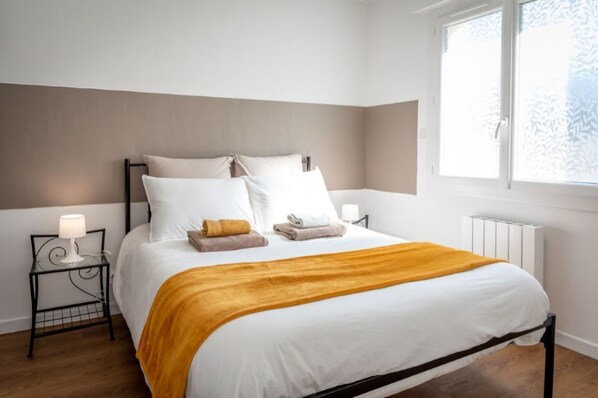 A neatly made bed with white linens and a mustard-colored throw blanket, two bedside tables with lamps, and a window with frosted glass.