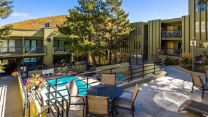 A pool area with outdoor seating, a building with multiple balconies, and trees in the background. - Summer at Resort - Pool, Hot Tub, Sauna, AC & Gym (Park City)