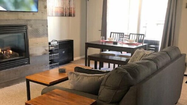 A living room with a gray sofa, a wooden coffee table, a fireplace, and a dining area with a table and chairs. - Summer at Resort - Pool, Hot Tub, Sauna, AC & Gym (Park City)