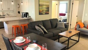A modern living room with a sofa, a dining table set for two, and a kitchen area in the background. - Summer at Resort - Pool, Hot Tub, Sauna, AC & Gym (Park City)