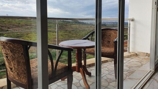 A balcony with a table and two chairs, overlooking a landscape with greenery and distant hills. - Greenzone BnB (Kiambu County)