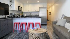Eine moderne Küche mit weißen Schränken, einer eingebauten Mikrowelle und einem Edelstahlkühlschrank. Vier rote Hocker stehen um einen Holztisch, und im Wohnbereich befindet sich eine graue Couch.