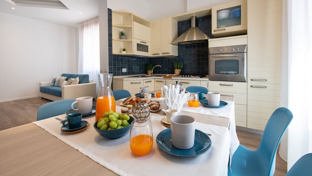 A modern kitchen with a dining area, featuring a breakfast setup with fruits, beverages, and a coffee table. - Nida 18 - Two Bedroom Apartment, Sleeps 6 (Cattolica)