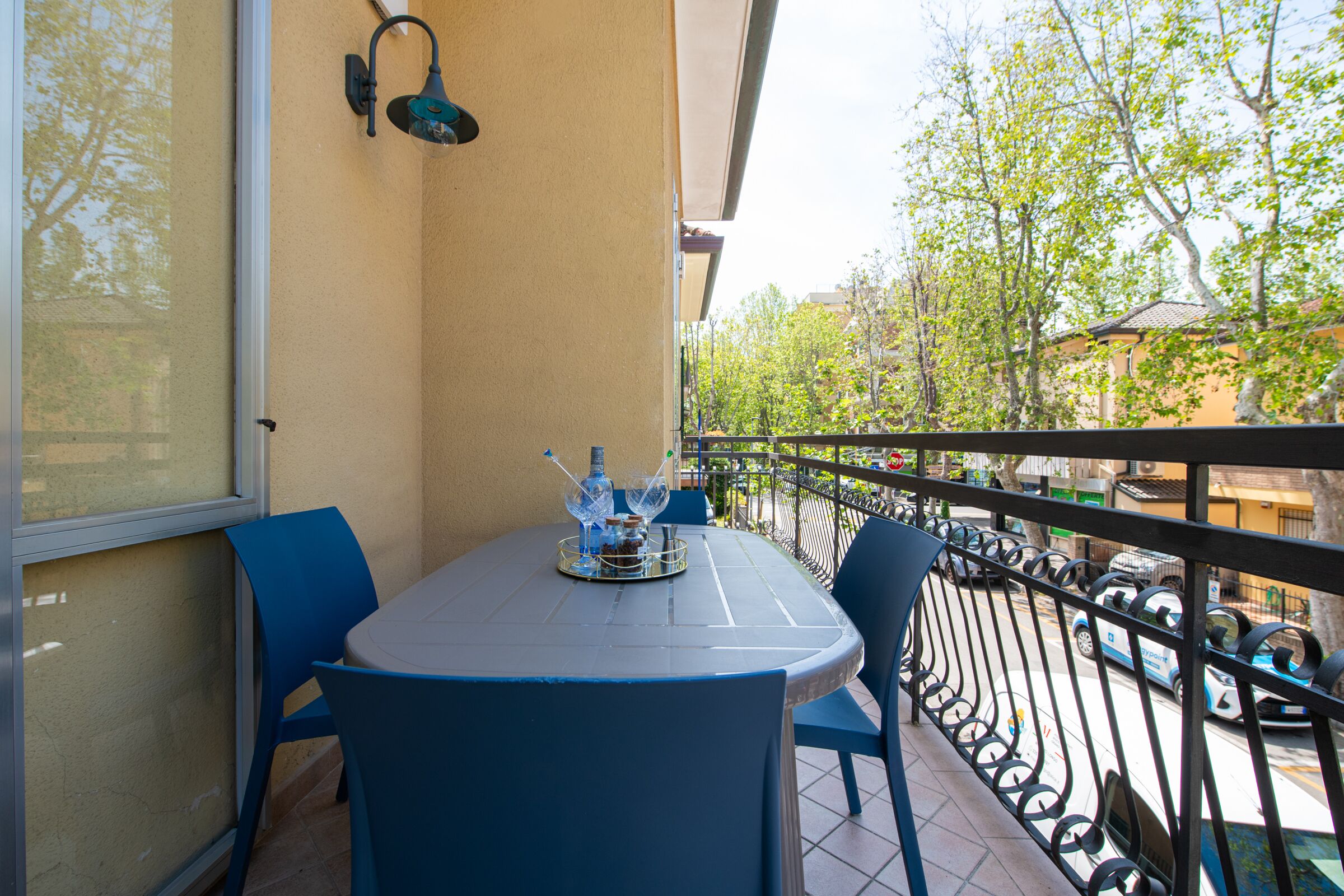 Een balkon met een tafel, stoelen, een raam en uitzicht op de straat en bomen.