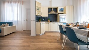 A modern kitchen with white cabinets, a stainless steel range hood, and a built-in oven. There is a dining area with a table and chairs, and a sofa in the adjacent living room. - Nida 18 - Two Bedroom Apartment, Sleeps 6 (Cattolica)