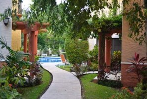 Ein Weg führt zu einem Poolbereich, der von Grünflächen und architektonischen Säulen umgeben ist.