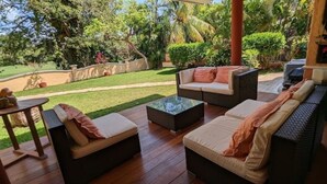 Un patio al aire libre con mobiliario de mimbre, una mesa de centro de vidrio y vistas a un césped y árboles bien cuidados.