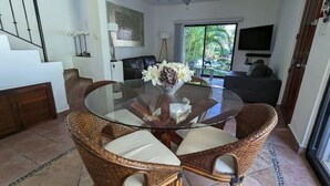 Un comedor con una mesa de vidrio rodeada de sillas de mimbre, un florero con flores y vistas a una sala de estar con un sofá y un televisor.