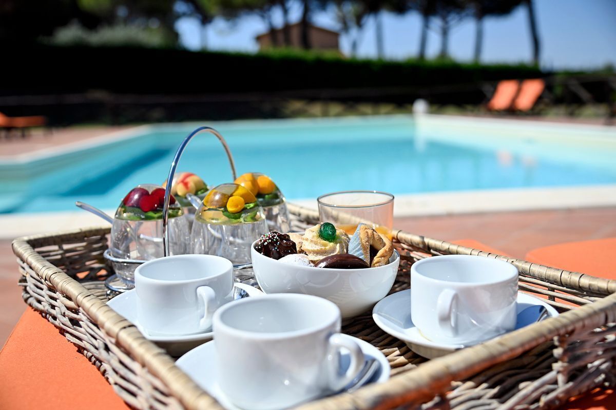 A tray with cups, a glass, and a bowl of assorted fruits and pastries by a pool.