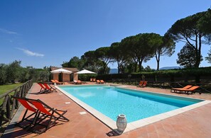 Une piscine rectangulaire bordée d’une terrasse carrelée, entourée de chaises longues orange et de parasols, avec des arbres et un ciel dégagé en arrière-plan.