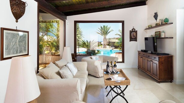 A spacious living room with a beige sofa, a coffee table with bottles, and a television on a wooden cabinet. A large window offers a view of a swimming pool and palm trees. - Camilla Canna - Three Bedroom Villa, Sleeps 6 (Cala Rossa)
