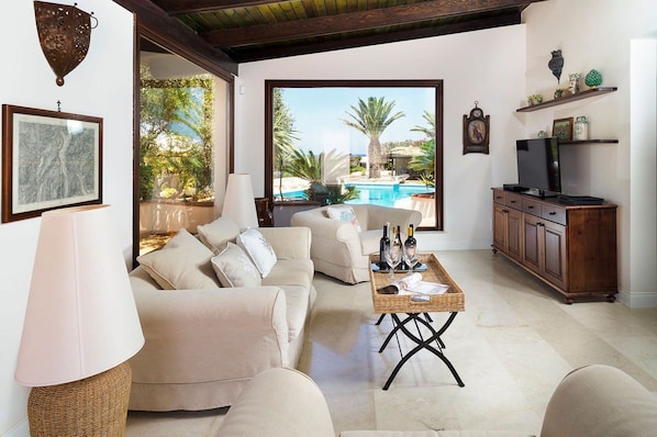 Ein geräumiges Wohnzimmer mit einem beigen Sofa, einem Couchtisch mit Flaschen und einem Fernseher auf einem Holzschrank. Ein großes Fenster bietet Blick auf einen Swimmingpool und Palmen.