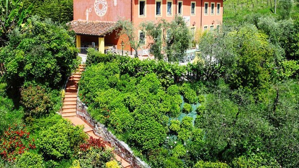 A large, two-story building with a red facade and yellow trim, surrounded by lush greenery and a small stream. - Dimora Dayan - Nine Bedroom Villa, Sleeps 18 (Capannori)