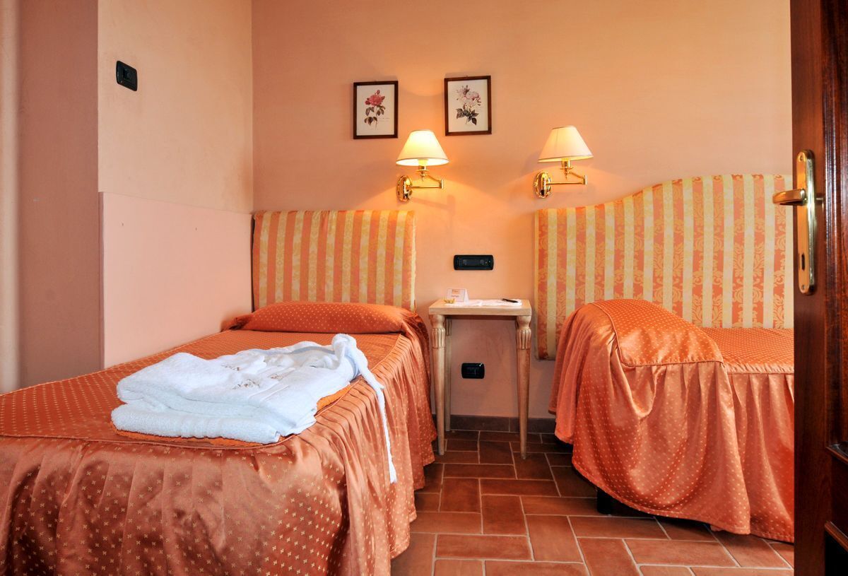 Une chambre d’hôtel avec deux lits, chacun recouvert d’une couverture orange et agrémenté d’une serviette blanche. Deux lampes murales sont fixées de chaque côté du lit. La chambre a un sol carrelé et les murs sont recouverts d’un papier peint à rayures.