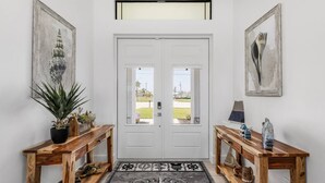 A hallway with a white door, two wooden side tables with decorative items, a black and white patterned rug, and framed artwork on the walls.