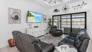 A spacious living room with a large flat-screen TV, leather sofas, a ceiling fan, and a view of the outdoors through large sliding glass doors.
