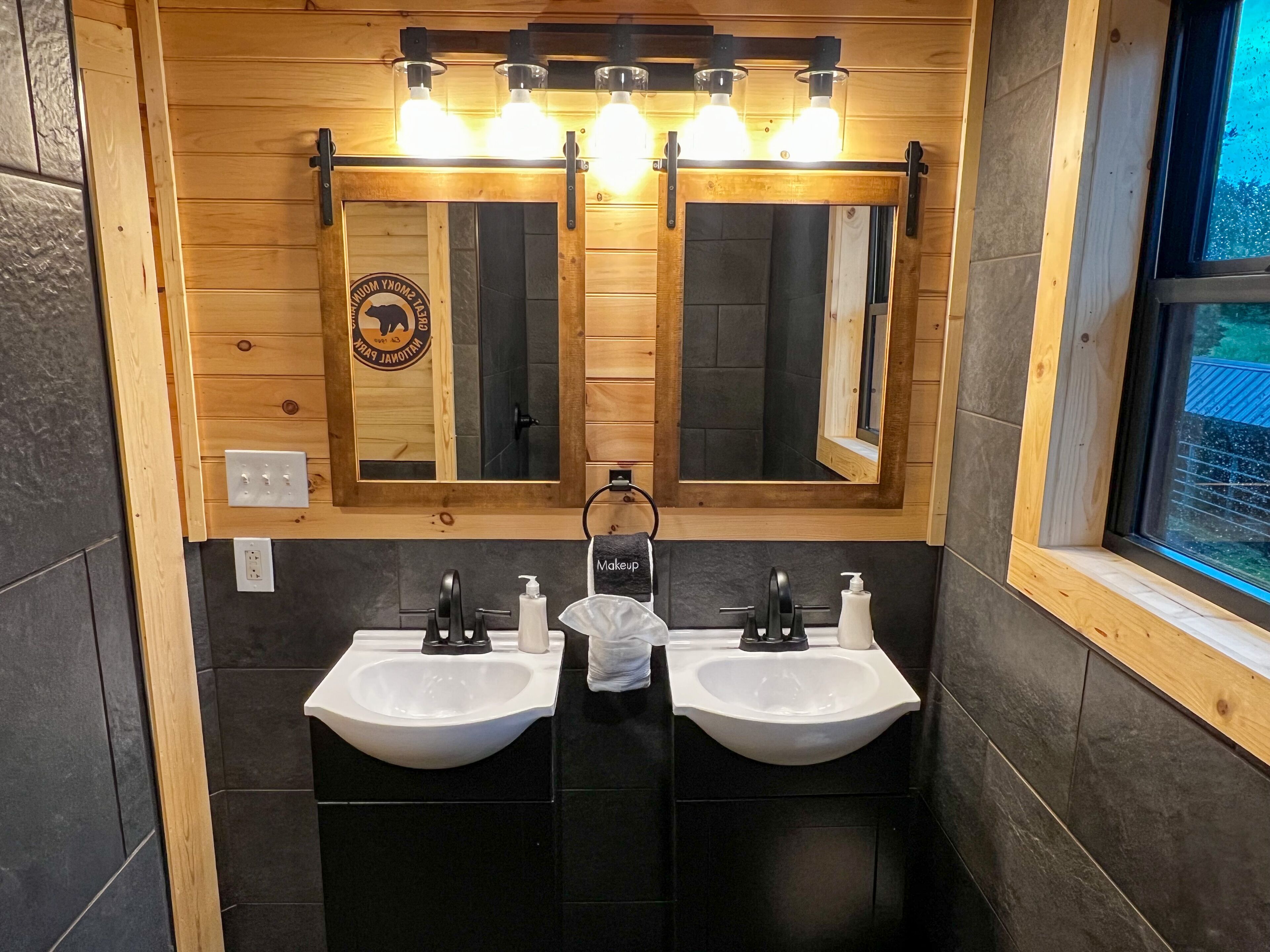 A bathroom with two sinks, wooden walls, a large mirror, and a window.