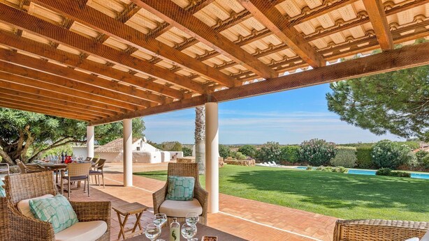 Une terrasse extérieure aménagée avec des meubles en osier, une table avec des verres et une vue sur une piscine et un jardin.