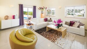 Un salon moderne avec un canapé blanc, un fauteuil jaune, une table basse et une vue sur une piscine, visible à travers de grandes fenêtres.