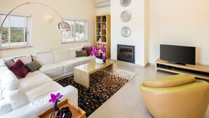 Un salon moderne avec un canapé blanc, une table basse, un pouf jaune, un téléviseur à écran plat et une cheminée.