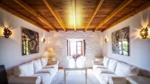 Una habitación con pared de piedra, vigas de madera en el techo, sofás blancos, una mesa central e iluminación instalada en la pared.