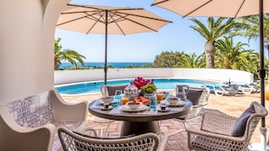 A patio with a pool, wicker chairs, and a table set with fruits and drinks, overlooking a clear blue sea. - Villa Lua - Three Bedroom Villa, Sleeps 6 (Lagoa)