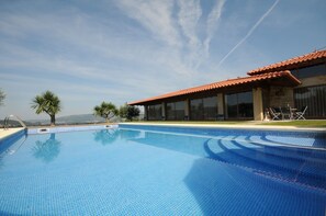 A swimming pool with clear blue water, a tiled deck, and a building with a red-tiled roof and large windows.