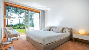 A modern bedroom with a large bed, a chair, a bedside table with a lamp, and a view of the outdoors through a large window. - Villa Senande - Five Bedroom Villa, Sleeps 10 (Âncora)