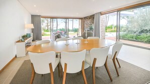 A modern dining area with a round table and six chairs, a small side table with a lamp, and a view of a garden through large windows. - Villa Senande - Five Bedroom Villa, Sleeps 10 (Âncora)