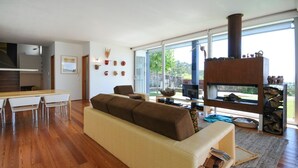 A modern living room with a sofa, a dining table with chairs, a fireplace, and a large window offering a view of the outdoors. - Moradia Moledo - Three Bedroom Villa, Sleeps 6 (Moledo)