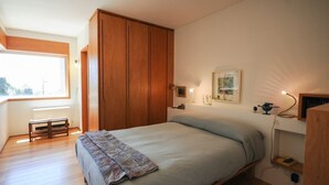 A bedroom with a bed, wooden wardrobe, bedside table, lamp, and a window. - Moradia Moledo - Three Bedroom Villa, Sleeps 6 (Moledo)