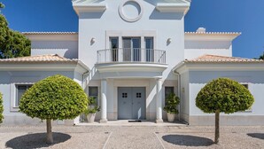 Un edificio de dos pisos con fachada blanca, un balcón y dos árboles simétricos en la entrada.