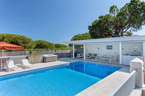 Une piscine rectangulaire bordée de carreaux, entourée d’une clôture blanche. On y trouve des chaises longues et un espace abrité avec un parasol rouge.