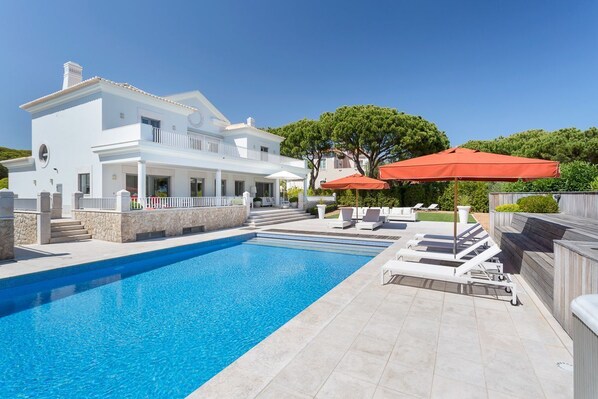Une piscine avec des chaises longues et des parasols, un bâtiment blanc doté d’un balcon, et un ciel d’un bleu limpide.