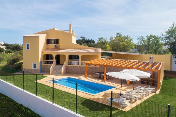 Una casa amarilla de dos pisos con piscina, mobiliario exterior y una pérgola.