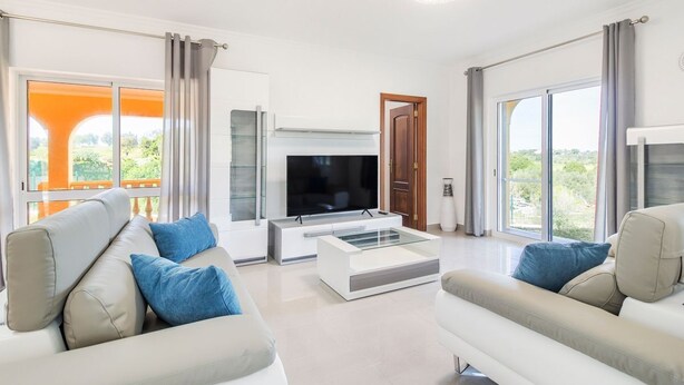 A modern living room with a flat-screen TV, two sofas, a coffee table, and large windows offering a view of greenery. - Villa Botela - Three Bedroom Villa, Sleeps 6 (Lagoa)