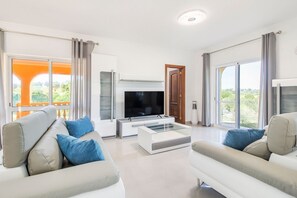 Una sala moderna con una televisión de pantalla plana, dos sofás, una mesa de centro y ventanas amplias que dan a vistas de vegetación.