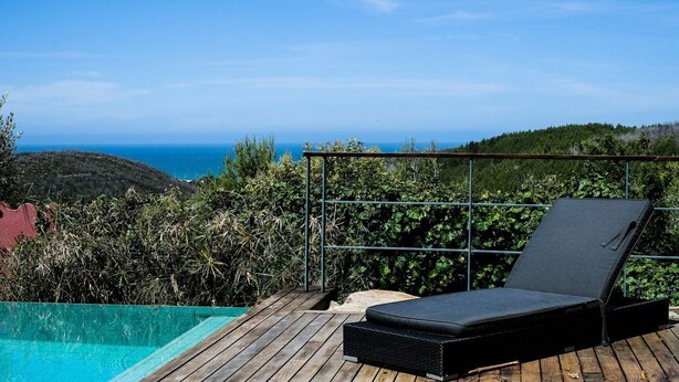 Una terraza de madera con una silla de descanso y una piscina, con vistas a un paisaje montañoso y al mar.