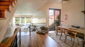 Amplia sala de estar con escalera de madera, mesa de comedor con sillas, ventana grande con vista a follaje y lámpara colgante.
