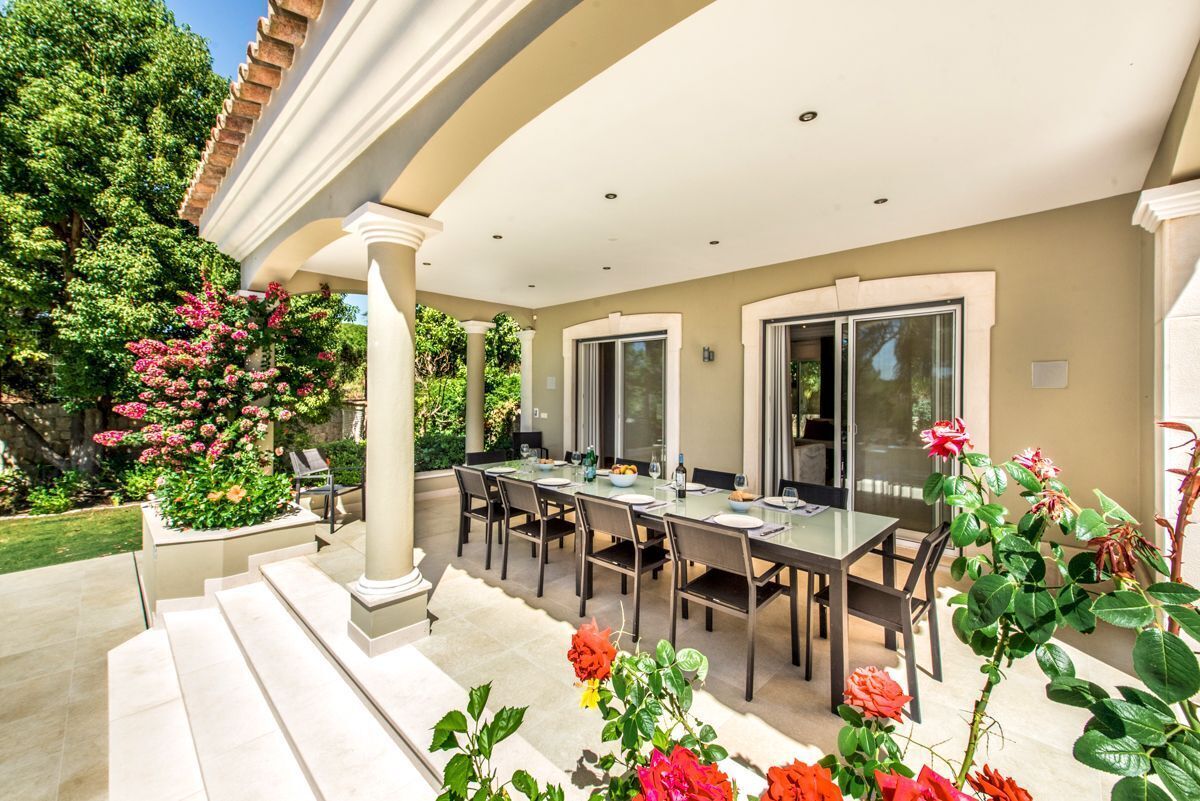 Um pátio externo com mesa e cadeiras de jantar, rodeado por vegetação exuberante e plantas floridas.